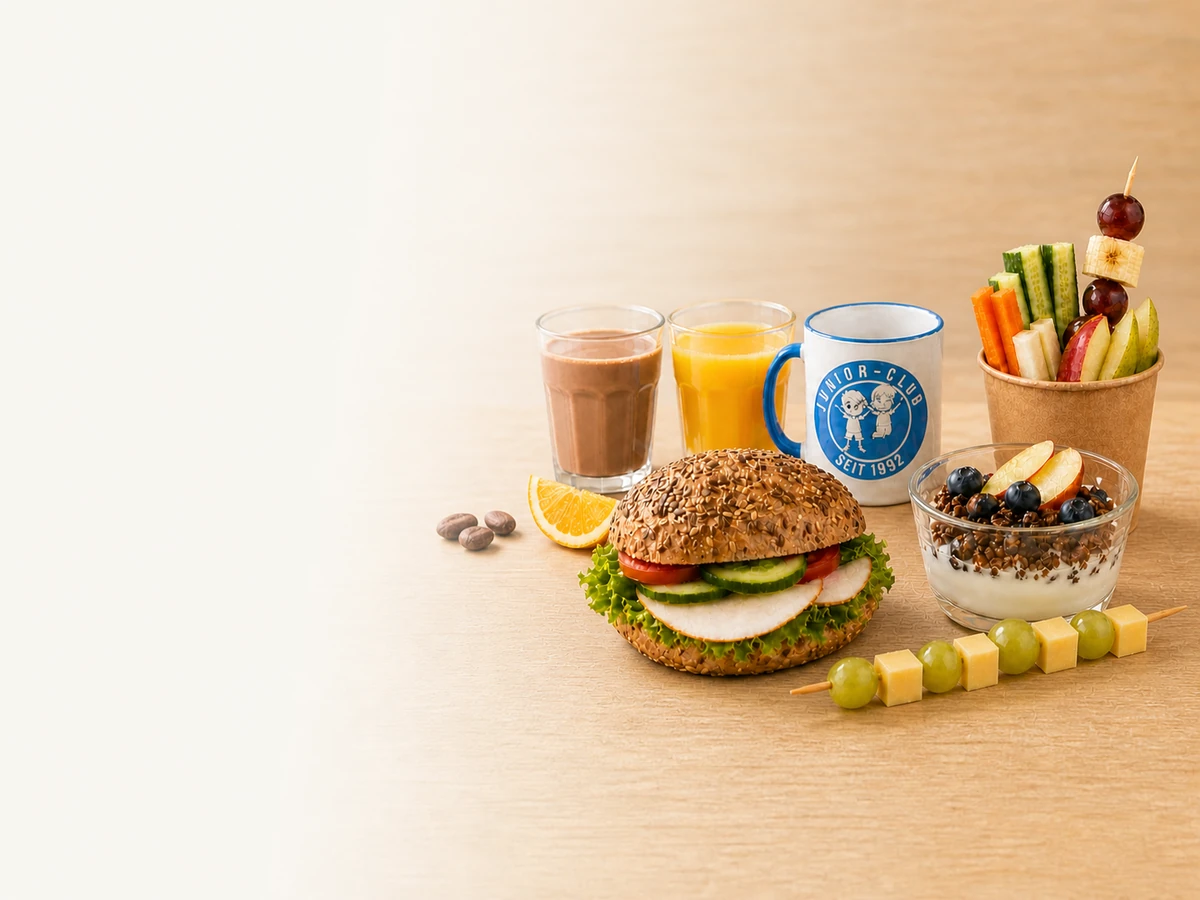 Belegtes Brötchen, Kakao und Orangensaft, ein Becher Joghurt mit Müsli und Obst, ein Obst-/Gemüsebecher mit Spieß und ein Käse-Trauben-Spieß, dekorativ mit einer Junior-Club-Tasse arrangiert